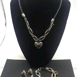 K&ouml;nnte beinhalten: Ein Schmuckset mit Halskette, Ohrringen und Armband. Die Halskette hat ein schwarzes geflochtenes Lederband und eine silber- und goldfarbene Kette mit Herzanh&auml;nger. Die Ohrringe und das Armband haben passende Herz- und Kettenmuster.