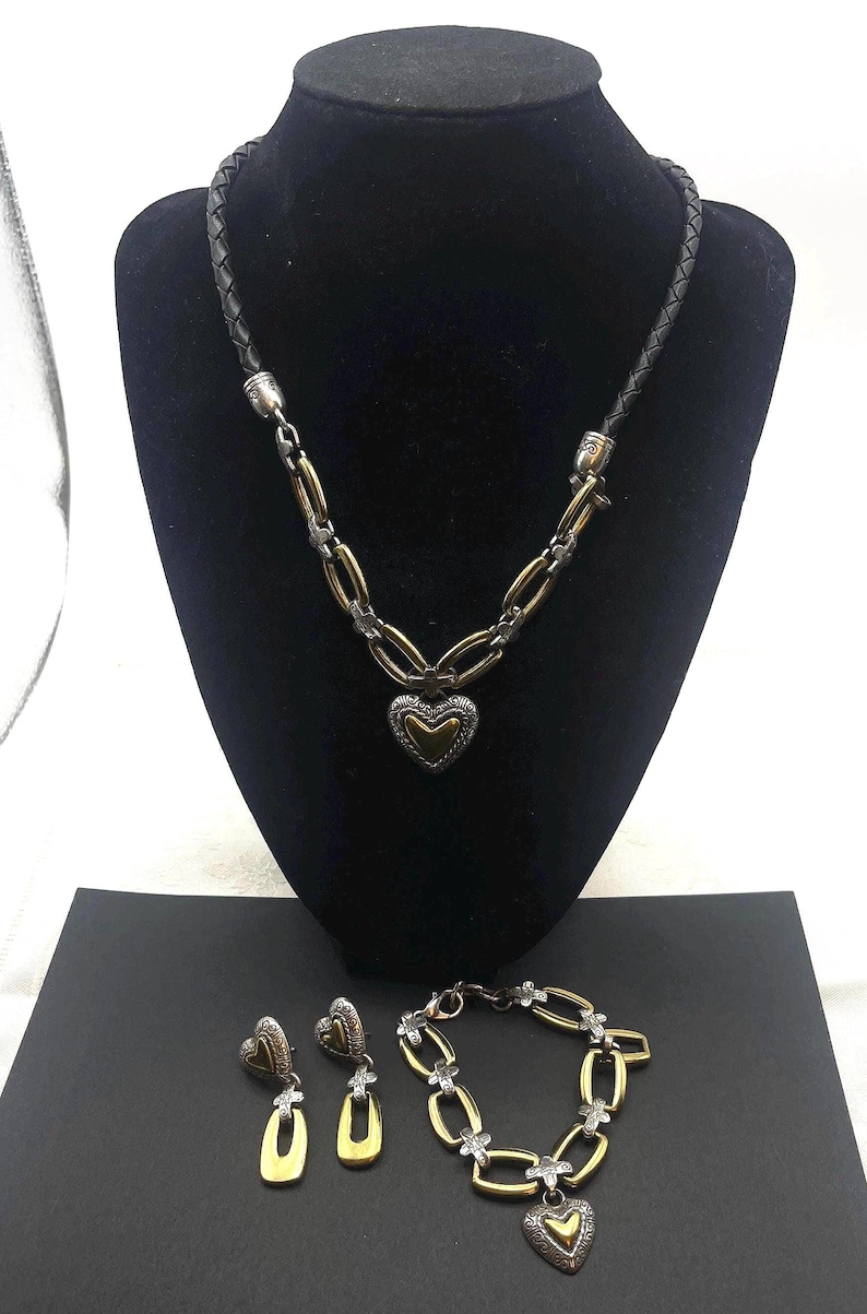 K&ouml;nnte beinhalten: Ein Schmuckset mit Halskette, Ohrringen und Armband. Die Halskette hat ein schwarzes geflochtenes Lederband und eine silber- und goldfarbene Kette mit Herzanh&auml;nger. Die Ohrringe und das Armband haben passende Herz- und Kettenmuster.