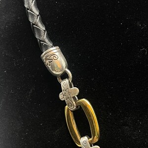 K&ouml;nnte beinhalten: Nahaufnahme einer Halskette mit geflochtener schwarzer Lederschnur. Die Halskette hat silberfarbene Metallakzente mit Kreuzdesign und goldfarbene rechteckige Glieder. Der Verschluss und die Glieder sind aufwendig detailliert.