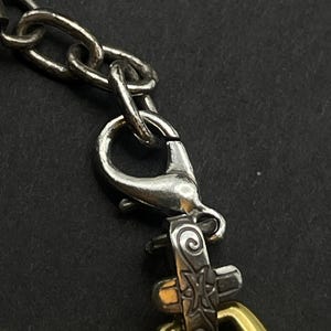 K&ouml;nnte beinhalten: Nahaufnahme einer silbernen Kette mit einem Karabinerverschluss und einem dekorativen goldfarbenen ovalen Glied. Der Verschluss und die Kette sind mit einem dekorativen Silberteil mit graviertem Design verbunden.