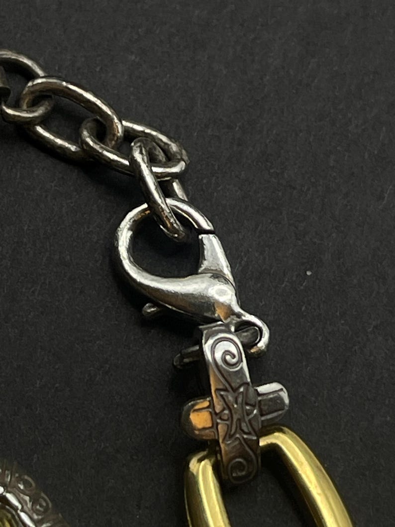 K&ouml;nnte beinhalten: Nahaufnahme einer silbernen Kette mit einem Karabinerverschluss und einem dekorativen goldfarbenen ovalen Glied. Der Verschluss und die Kette sind mit einem dekorativen Silberteil mit graviertem Design verbunden.