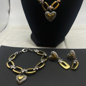 K&ouml;nnte beinhalten: Ein goldfarbenes Halsketten-, Armband- und Ohrring-Set. Der Schmuck hat silberfarbene Akzente und herzf&ouml;rmige Anh&auml;nger. Die Halskette und das Armband haben ein Kettenglied-Design. Die Ohrringe sind herzf&ouml;rmig mit Gold- und Silber-Finish.