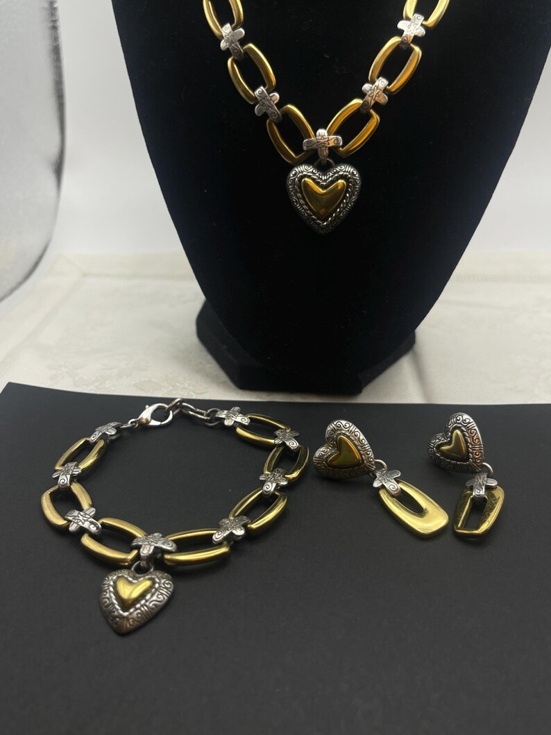 K&ouml;nnte beinhalten: Ein goldfarbenes Halsketten-, Armband- und Ohrring-Set. Der Schmuck hat silberfarbene Akzente und herzf&ouml;rmige Anh&auml;nger. Die Halskette und das Armband haben ein Kettenglied-Design. Die Ohrringe sind herzf&ouml;rmig mit Gold- und Silber-Finish.