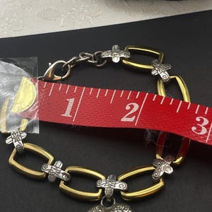 K&ouml;nnte beinhalten: Ein gold- und silberfarbenes Armband mit Herzanh&auml;nger. Das Armband ist etwa 6,35 cm lang. Es hat einen silberfarbenen Verschluss und silberfarbene Verzierungen auf den goldfarbenen Gliedern.