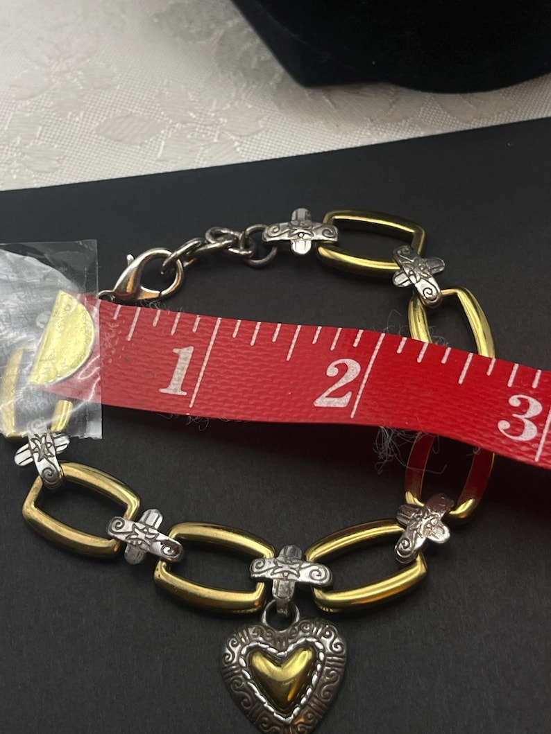 K&ouml;nnte beinhalten: Ein gold- und silberfarbenes Armband mit Herzanh&auml;nger. Das Armband ist etwa 6,35 cm lang. Es hat einen silberfarbenen Verschluss und silberfarbene Verzierungen auf den goldfarbenen Gliedern.