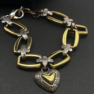 K&ouml;nnte beinhalten: Ein gold- und silberfarbenes Armband mit Herzanh&auml;nger. Das Armband hat goldfarbene rechteckige Glieder, silberfarbene kreuzf&ouml;rmige Akzente und einen herzf&ouml;rmigen Anh&auml;nger mit goldenem Zentrum und aufwendigen silbernen Details. Der Verschluss ist silberfarben.