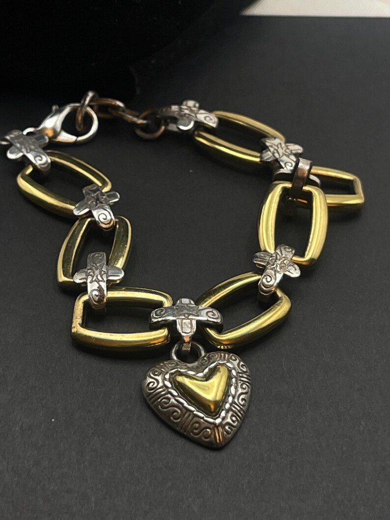 K&ouml;nnte beinhalten: Ein gold- und silberfarbenes Armband mit Herzanh&auml;nger. Das Armband hat goldfarbene rechteckige Glieder, silberfarbene kreuzf&ouml;rmige Akzente und einen herzf&ouml;rmigen Anh&auml;nger mit goldenem Zentrum und aufwendigen silbernen Details. Der Verschluss ist silberfarben.
