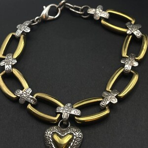 K&ouml;nnte beinhalten: Ein silber- und goldfarbenes Armband mit herzf&ouml;rmigem Anh&auml;nger. Das Armband hat ovale goldene Glieder, die durch silberne kreuzf&ouml;rmige Akzente verbunden sind. Der Herzanh&auml;nger hat eine goldene Mitte und einen verzierten silbernen Rand.