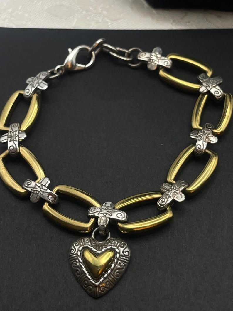 K&ouml;nnte beinhalten: Ein silber- und goldfarbenes Armband mit herzf&ouml;rmigem Anh&auml;nger. Das Armband hat ovale goldene Glieder, die durch silberne kreuzf&ouml;rmige Akzente verbunden sind. Der Herzanh&auml;nger hat eine goldene Mitte und einen verzierten silbernen Rand.
