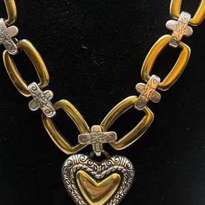 K&ouml;nnte beinhalten: Eine gold- und silberfarbene Halskette mit Herzanh&auml;nger. Die Halskette hat eine Kette aus goldfarbenen rechteckigen Gliedern, die durch silberfarbene kreuzf&ouml;rmige Akzente verbunden sind. Der Herzanh&auml;nger ist goldfarben mit einem silbernen Filigranrand.