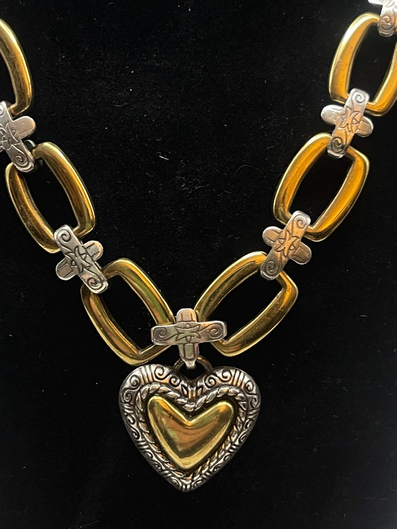K&ouml;nnte beinhalten: Eine gold- und silberfarbene Halskette mit Herzanh&auml;nger. Die Halskette hat eine Kette aus goldfarbenen rechteckigen Gliedern, die durch silberfarbene kreuzf&ouml;rmige Akzente verbunden sind. Der Herzanh&auml;nger ist goldfarben mit einem silbernen Filigranrand.