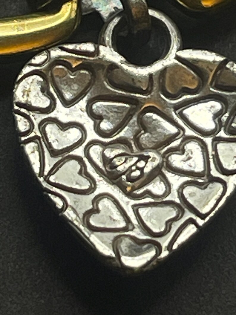 K&ouml;nnte beinhalten: Nahaufnahme eines silbernen herzf&ouml;rmigen Anh&auml;ngers mit einem Muster aus kleinen Herzen. Der Anh&auml;nger hat eine strukturierte Oberfl&auml;che und eine Schlaufe oben zum Befestigen an einer Kette. Das Metall hat ein angelaufenes Aussehen.