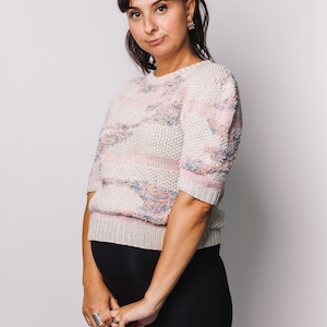 May include: A knitted sweater with short sleeves, featuring a cream base with pink, blue, and yellow accents. The sweater is paired with black leggings. The person in the image is wearing a black bow and earrings.