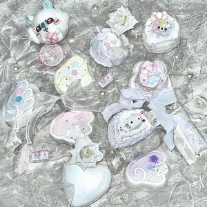 Könnte beinhalten: Eine Auswahl pastellfarbener Dekorationsartikel, darunter Plüschtiere, herzförmige Ornamente und kleine, runde Schmuckstücke. Die Artikel sind mit Schleifen, Spitze und floralen Akzenten verziert und auf einer schimmernden, silbernen Oberfläche angeordnet. Weiße Rosen und Kunstperlen sind verstreut.