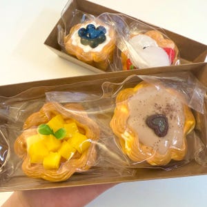May include: Four decorative pastries in a cardboard box, each individually wrapped in clear plastic. The pastries have various toppings, including blueberries, strawberries, mango, and a heart-shaped design. The pastries are golden brown with a layered texture.