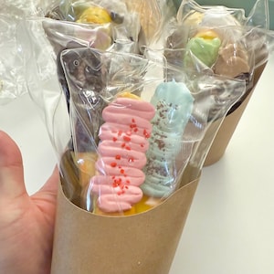Può includere: Una collezione di caramelle colorate di varie forme, confezionate singolarmente in plastica trasparente e presentate in un cono di carta marrone. Le caramelle includono tonalità rosa, blu e gialle, con zuccherini e altre decorazioni.