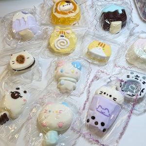 Puede incluir: Una variedad de postres individuales, coloridos y caprichosos, envueltos. Los dulces incluyen pasteles con forma de gato, barras de helado y un diseño de té boba. Los postres están en envases de plástico transparente con un borde de encaje decorativo.