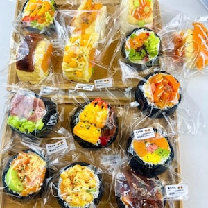 May include: A wooden tray displays various sushi rolls and sandwiches, each individually wrapped in clear plastic. The sushi features colourful fillings like orange, yellow, and green, while the sandwiches have a similar vibrant appearance. The food items are arranged neatly.