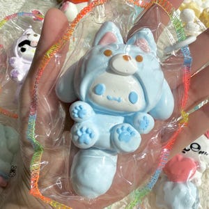Può includere: Una statuetta di un personaggio dei cartoni animati blu chiaro in un involucro di plastica trasparente. La statuetta ha un cappuccio a forma di coniglio con una faccia da orso, zampe blu e una faccia sorridente. Altre statuette sono visibili sullo sfondo.