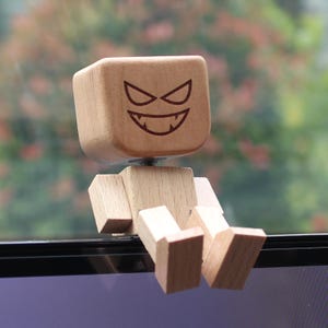 以下が含まれることがあります： 四角い頭部を持つ明るい茶色の木製フィギュア。レーザー刻印された顔には、鋭い目と歯を見せた大きな笑みが描かれています。ブロック状の胴体、長方形の腕、そして座った姿勢の脚部で構成されています。