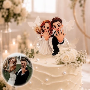 Op de afbeelding: Een taarttopper met twee chibi-figuren, één met lang roodbruin haar in een witte jurk en één met kort bruin haar in een zwart pak, die beide hun handen met ringen tonen. Inclusief witte vlinderaccenten en gedroogde bloemen.