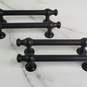 May include: Four black cabinet pulls with a classic design are arranged on a white marble surface. Each pull features a cylindrical bar with decorative end pieces and a pair of supporting feet.