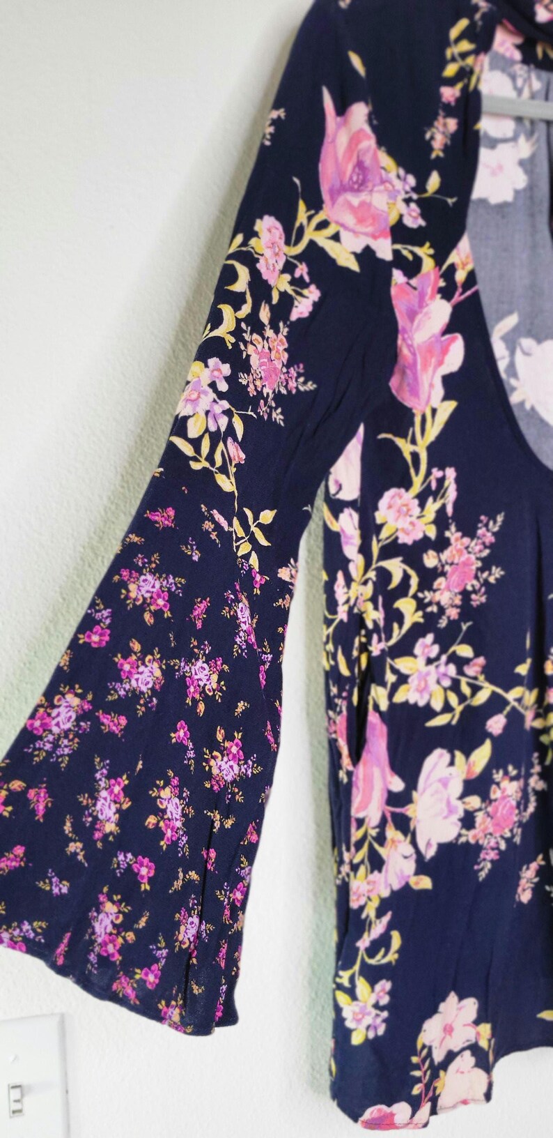May include: Navy blue top with a floral pattern in pink, purple, and yellow. The top has long, bell sleeves and a scoop neckline. The floral print is scattered across the fabric.