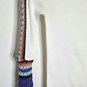 May include: White long-sleeved shirt with a textured waffle knit. The left sleeve features a colorful crochet design with orange, blue, and black accents. The sleeve's cuff has a blue and gray patterned design.