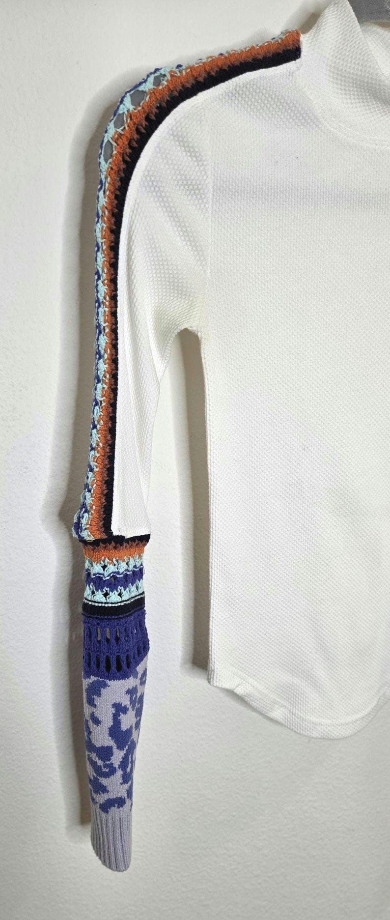 May include: White long-sleeved shirt with a textured waffle knit. The left sleeve features a colorful crochet design with orange, blue, and black accents. The sleeve's cuff has a blue and gray patterned design.