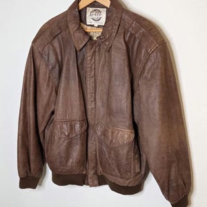 May include: Brown leather bomber jacket with two front pockets and a ribbed hem and cuffs. The jacket has a collared neckline and a label on the inside collar. The jacket is hanging on a wooden hanger.