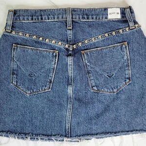 May include: A blue denim skirt with two back pockets and a raw hem. The waistband is embellished with silver-colored studs. The skirt has a classic five-pocket design and a button and zipper closure.