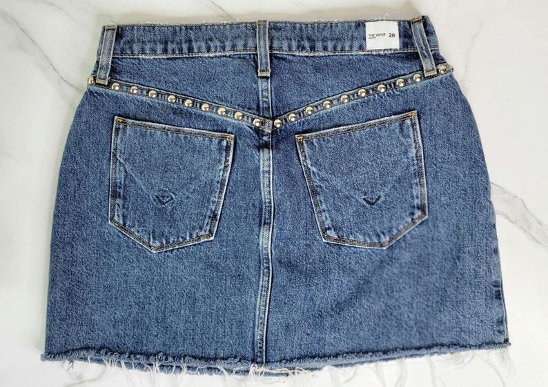 May include: A blue denim skirt with two back pockets and a raw hem. The waistband is embellished with silver-colored studs. The skirt has a classic five-pocket design and a button and zipper closure.