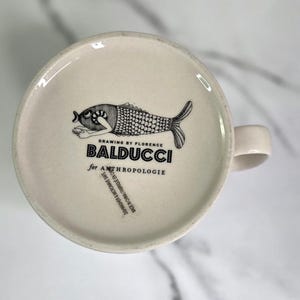 Puede incluir: Taza de cer&aacute;mica color crema con una ilustraci&oacute;n de pez en blanco y negro en la base. El texto "DRAWING BY FLORENCE BALDUCCI for ANTHROPOLOGIE" est&aacute; impreso debajo del pez. La taza tiene un asa curvada.