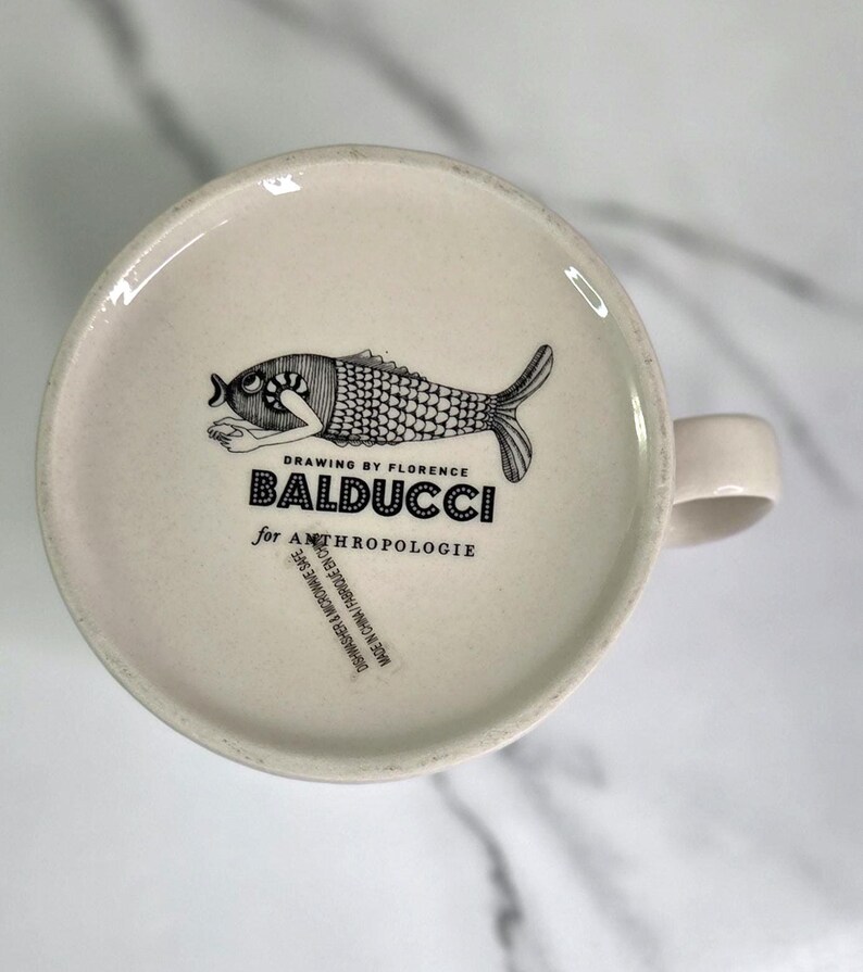 Puede incluir: Taza de cer&aacute;mica color crema con una ilustraci&oacute;n de pez en blanco y negro en la base. El texto "DRAWING BY FLORENCE BALDUCCI for ANTHROPOLOGIE" est&aacute; impreso debajo del pez. La taza tiene un asa curvada.