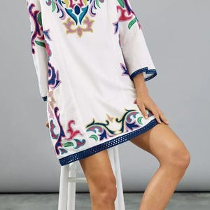 May include: White tunic dress with colorful embroidered floral designs. The dress features a V-neck, three-quarter sleeves, and a navy blue trim along the hem and sleeves. The embroidery includes pink, yellow, blue, and green accents. The model is wearing sandals.