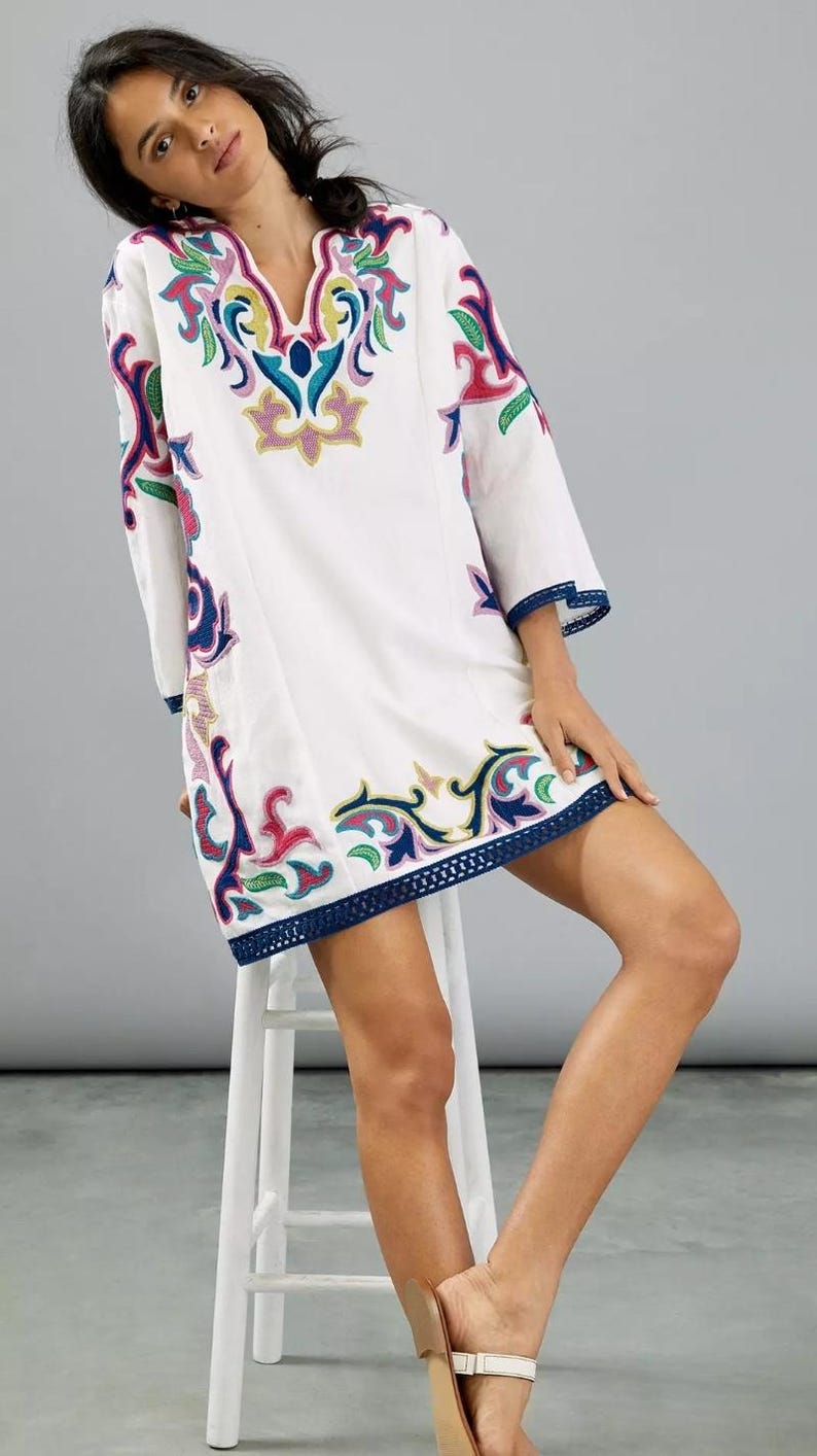 May include: White tunic dress with colorful embroidered floral designs. The dress features a V-neck, three-quarter sleeves, and a navy blue trim along the hem and sleeves. The embroidery includes pink, yellow, blue, and green accents. The model is wearing sandals.