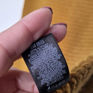 Może przedstawiać: Zbliżenie czarnej metki z białym tekstem, na tle musztardowoż&oacute;łtej dzianiny. Na metce widnieje napis "100% ACRYLIC" i instrukcje prania. Tkanina wydaje się być szalikiem lub podobnym przedmiotem.