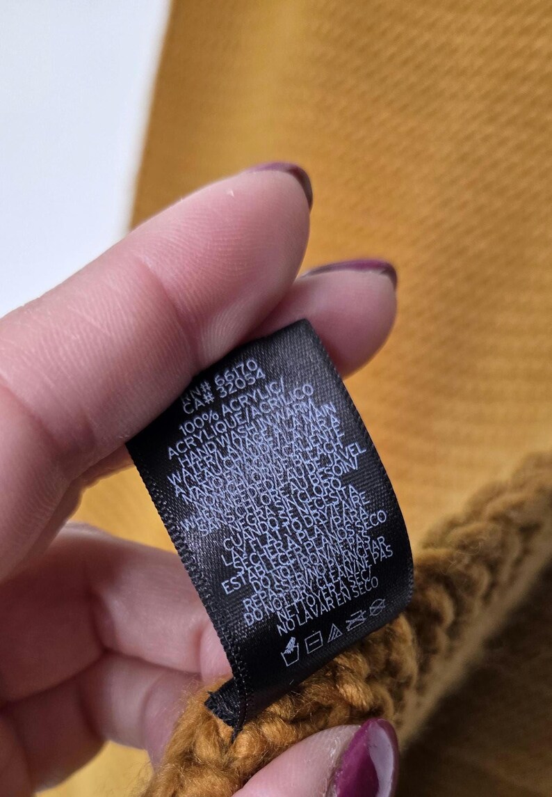 Może przedstawiać: Zbliżenie czarnej metki z białym tekstem, na tle musztardowoż&oacute;łtej dzianiny. Na metce widnieje napis "100% ACRYLIC" i instrukcje prania. Tkanina wydaje się być szalikiem lub podobnym przedmiotem.