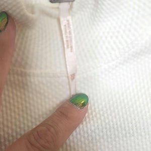 May include: Close-up of a cream-colored textured fabric. A small tag with the brand name is attached to the fabric. The fabric appears to be part of a garment, possibly a sweater or top.