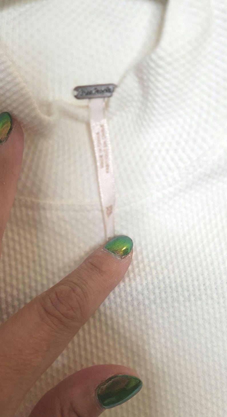 May include: Close-up of a cream-colored textured fabric. A small tag with the brand name is attached to the fabric. The fabric appears to be part of a garment, possibly a sweater or top.