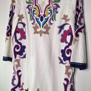 May include: White tunic with colorful embroidered floral design. The tunic has a V-neck and three-quarter sleeves. The embroidery features shades of pink, blue, green, and yellow. The sleeves and hem are trimmed with a navy blue border.