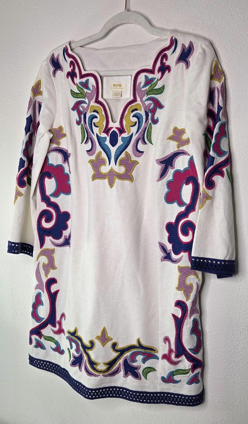 May include: White tunic with colorful embroidered floral design. The tunic has a V-neck and three-quarter sleeves. The embroidery features shades of pink, blue, green, and yellow. The sleeves and hem are trimmed with a navy blue border.