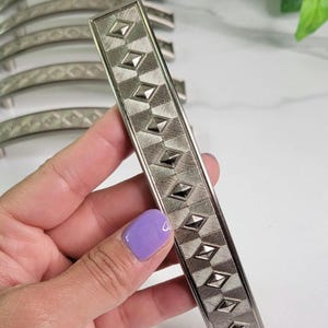 May include: A silver cabinet hardware pull with a geometric pattern. The pull is rectangular and has a slightly curved shape.