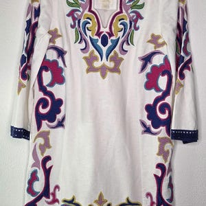 May include: White tunic dress with colorful embroidered floral and scrollwork designs. The dress features a V-neckline and three-quarter sleeves with navy blue trim. The embroidery includes shades of pink, purple, blue, green, and yellow.