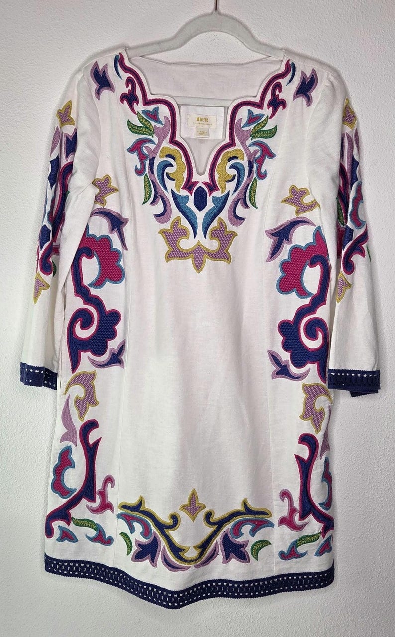 May include: White tunic dress with colorful embroidered floral and scrollwork designs. The dress features a V-neckline and three-quarter sleeves with navy blue trim. The embroidery includes shades of pink, purple, blue, green, and yellow.