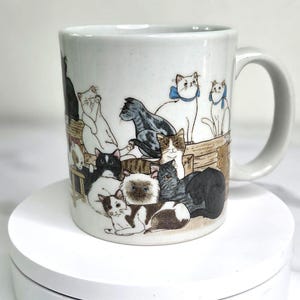 May include: White ceramic mug featuring a colorful illustration of various cats. The cats are depicted in different poses and colors, some with blue bows. The mug has a white handle and is set on a white surface.