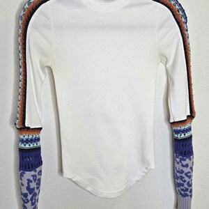 May include: White long-sleeve shirt with a mock turtleneck. The sleeves feature a unique design with crochet and patterned details in shades of blue, orange, and black. The shirt is hanging on a hanger against a white background.