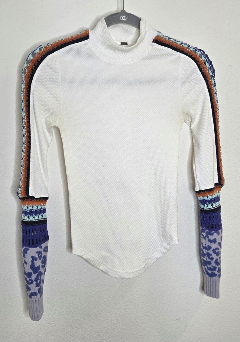 May include: White long-sleeve shirt with a mock turtleneck. The sleeves feature a unique design with crochet and patterned details in shades of blue, orange, and black. The shirt is hanging on a hanger against a white background.