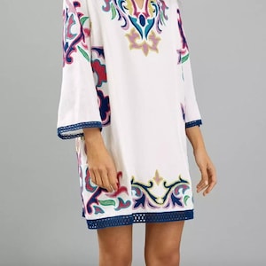 May include: White tunic dress with colorful embroidered floral patterns in pink, blue, green, and yellow. The dress features a V-neck and three-quarter sleeves with a blue trim. The hemline also has a blue trim.