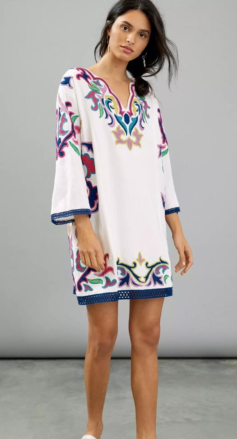 May include: White tunic dress with colorful embroidered floral patterns in pink, blue, green, and yellow. The dress features a V-neck and three-quarter sleeves with a blue trim. The hemline also has a blue trim.